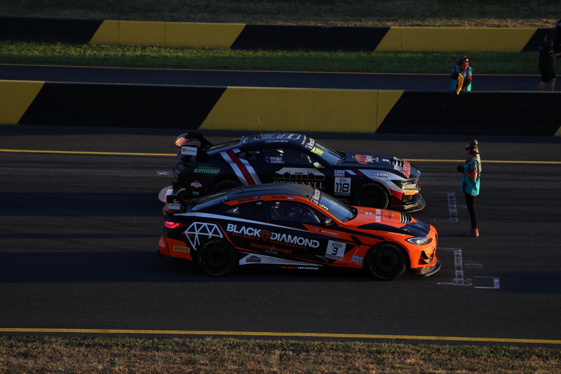 Orange and black BMW race car beside a black Mustang on the starting grid